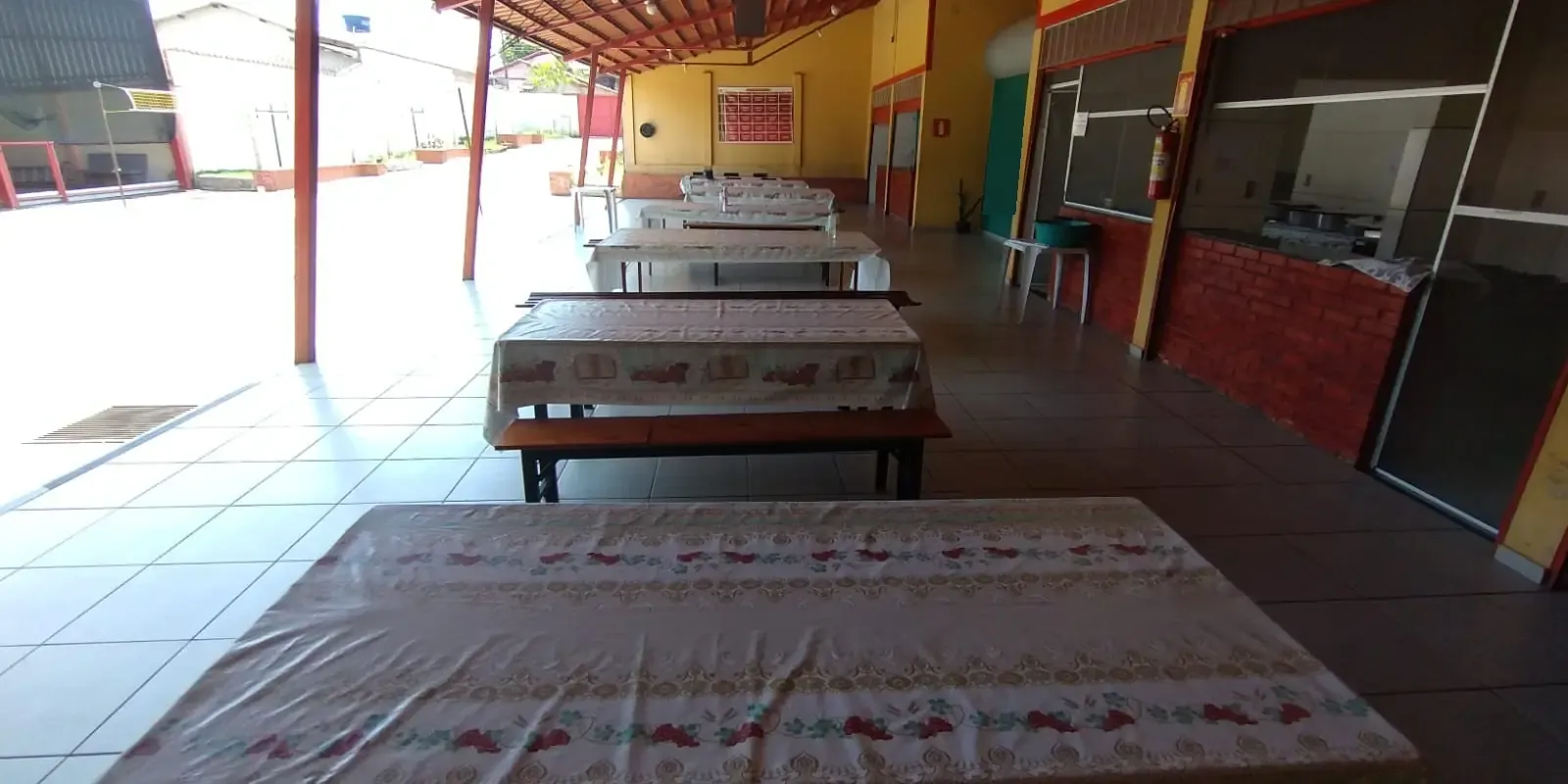 Refeitório coberto com mesas compridas, toalhas estampadas e bancos de madeira, aberto para um pátio ensolarado